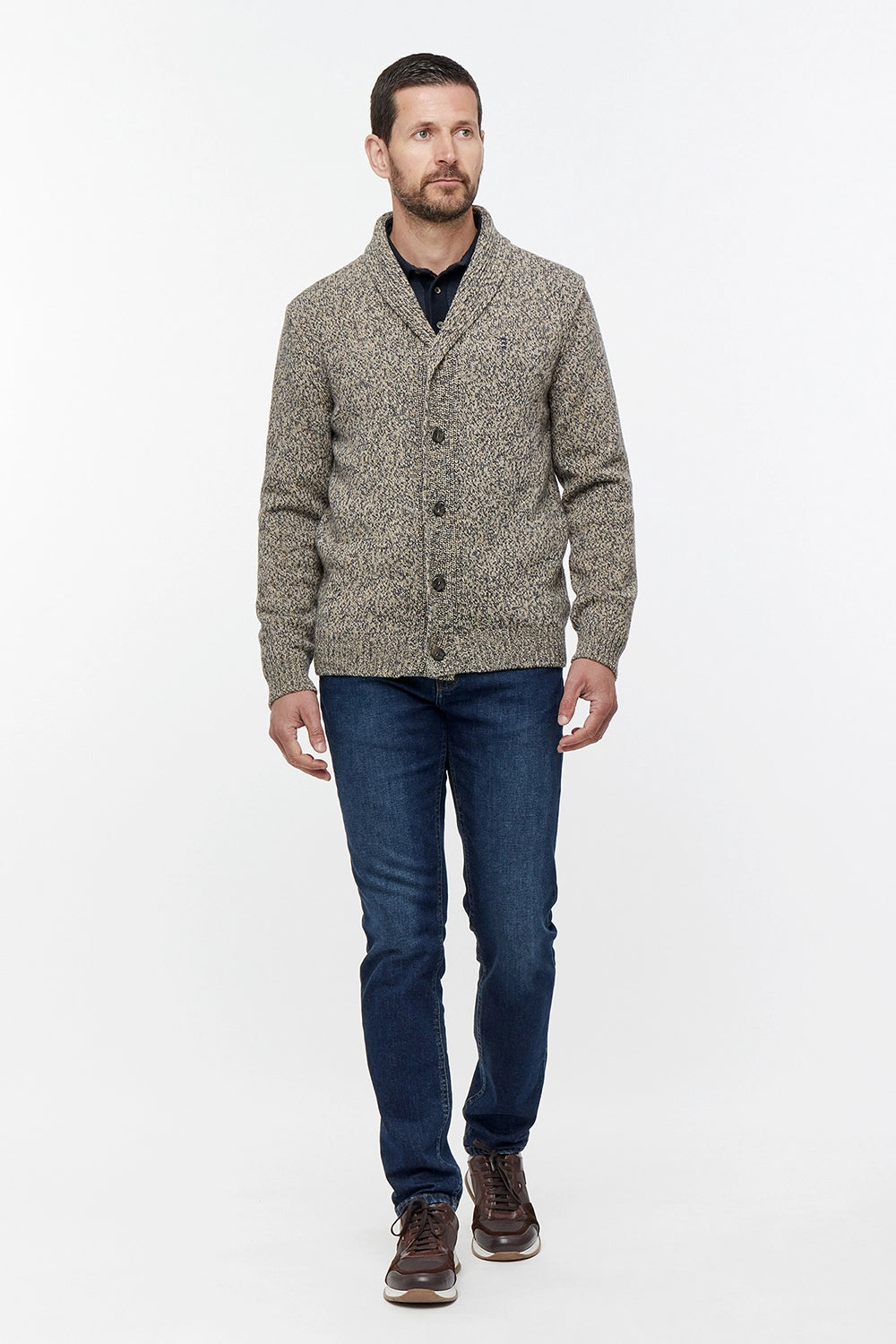 Man wearing a patterned cardigan, dark jeans, and brown shoes on a white background